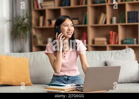 Positive koreanische Dame mit Telefongespräch, während auf der Couch zu Hause sitzen und mit Laptop, Kopierer Platz Stockfoto