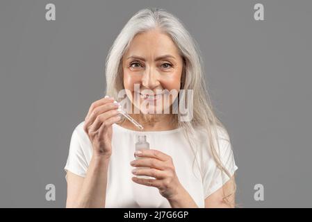 Hautpflege und Wellness. Porträt einer lächelnden älteren Frau, die Gesichtsserum mit Tropf auf grauem Studiohintergrund auftrage Stockfoto