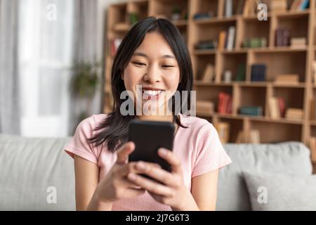 Positive koreanische Dame mit Smartphone, Surfen im Internet, Online-Chat, Video ansehen, zu Hause auf der Couch sitzen Stockfoto