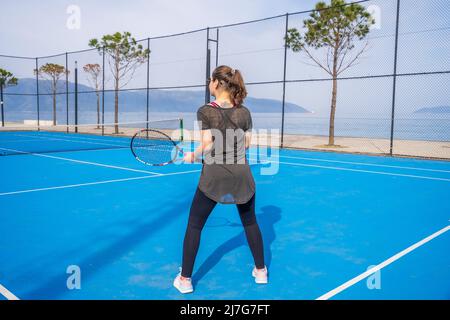 Das junge Mädchen in einem dunkelblauen Kleid spielt Tennis auf dem Platz mit einer harten blauen Beschichtung Stockfoto