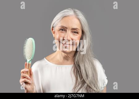 Anmutig altern. Charmante ältere Dame mit seidig langen Haaren, die Bürste hält und auf grauem Studiohintergrund lächelt Stockfoto