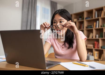 Unglaublich. Schockierte asiatische Dame in einer Brille, die auf den Bildschirm des Laptops schaute und zu Hause am Schreibtisch saß Stockfoto