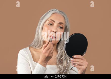 Schöne reife Dame, die Lippenstift vor dem Spiegel anwendet und sich auf braunem Studiohintergrund um sich selbst kümmert Stockfoto