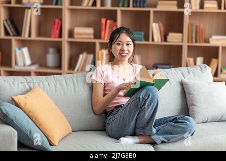 Konzept des häuslichen Lebens. Positive asiatische Dame, die Papierbuch hält, allein zu Hause auf der bequemen Couch sitzt, freier Platz Stockfoto