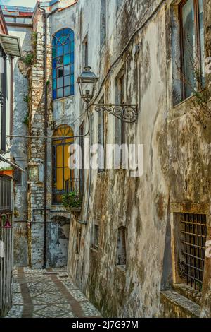 Charakteristische schmale Gasse zwischen hohen Gebäuden mit orangefarbenen und blauen Buntglasfenstern. Vico del Gargano, Provinz Foggia, Apulien, Italien Stockfoto