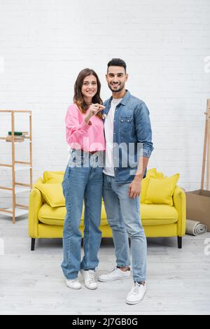 Positives multiethnisches Paar, das den Schlüssel hält und zu Hause auf die Kamera schaut Stockfoto