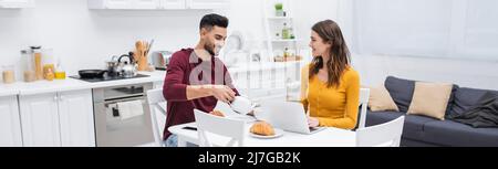 Positiver muslimischer Mann goss Tee neben meiner Freundin mit Laptop und Frühstück auf dem Tisch in der Küche, Banner Stockfoto