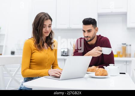 Lächelnde Frau mit Laptop in der Nähe von muslimischem Mann und Frühstück in der Küche Stockfoto