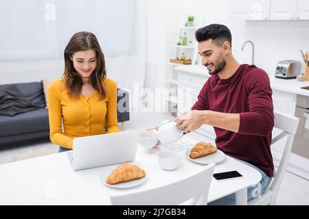 Fröhliches interrassisches Paar mit Laptop in der Nähe des Frühstücks in der Küche Stockfoto