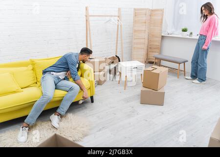 Junges interrassisches Paar, das in der Nähe von Paketen im Wohnzimmer eine bengalkatze ansieht Stockfoto