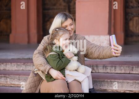 Lachendes Mädchen, das Teddybär in der Nähe der Mutter hält und Selfie auf dem Smartphone im Freien nimmt