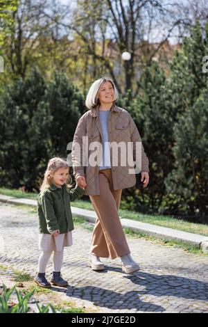 Volle Länge der glücklichen und stilvollen Frau mit Tochter zu Fuß im Frühling Park Stockfoto