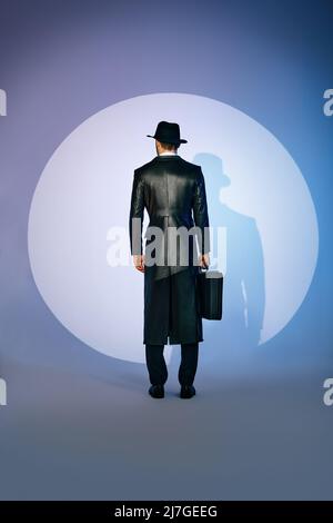 Rückansicht der Mann Silhouette in schwarzem Mantel und Hut hält Aktentasche im Rampenlicht auf Studio-Hintergrund. noir Film-Stil. Privatdetektiv, Spion, in Stockfoto