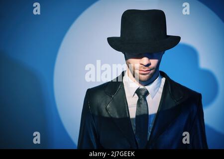 Porträt eines gutaussehenden Mannes im schwarzen Mantel, der sich hinter seinem Hut versteckt, im Rampenlicht auf dem Hintergrund des Studios. noir-Filmstil. Privatdetektiv, investiga Stockfoto