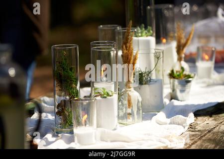 Hochzeitsfeier Bankett im Freien im Wald. Esstisch im Boho-Stil mit Kerzen, weißem Tuch, Blumen, serviert mit Tellern, Geschirr dekoriert Stockfoto