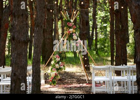 Wunderschöne böhmische Tipi-Bogendekoration auf der Hochzeitsfeier im Freien im Pinienwald mit Kegeln. Stühle, floristische Blumenkompositionen aus Rosen Stockfoto