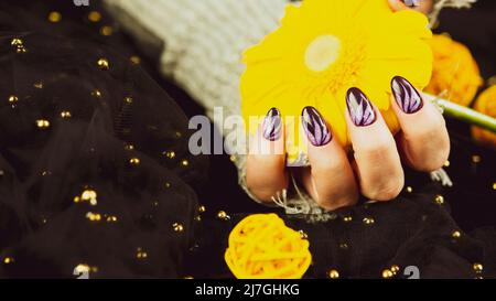Erwachsene Frauenhand in stylischem Strickhandschuh mit lila Maniküre auf schwarzem Hintergrund. Schneiden Sie nicht erkennbare Person mit Design auf den Nägeln. Konzept der Pflege Stockfoto