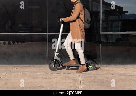 Unerkennbarer Geschäftsmann auf dem täglichen Pendelweg, der Mikroscooter fährt, der ein elegantes und modernes Outfit in braunen und beigen Farben mit Rucksack im b Stockfoto