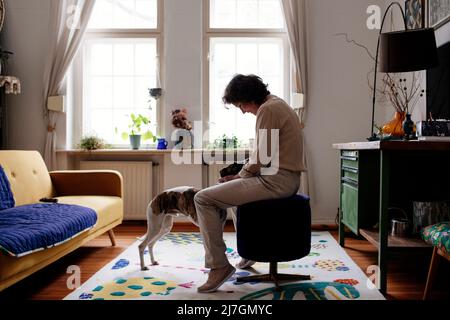 Seitenansicht einer älteren Frau, die den Hund streichelte, während sie auf einem Hocker im Wohnzimmer zu Hause saß Stockfoto