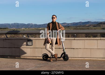 Junger Geschäftsmann auf seiner Arbeitspause für einen nachhaltigen und gesunden Lebensstil mit Elektroroller und recycelbarer Kaffeetasse Stockfoto