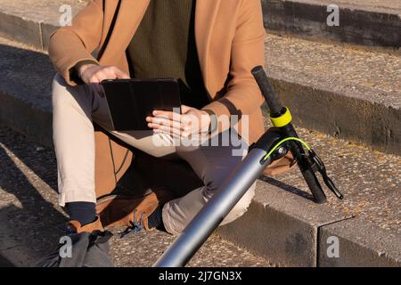 Beschnittene Nahaufnahme eines unkenntlichen Mannes, der mit Tablet und Elektroroller im Stadtpark sitzt und den sonnigen Tag im Sommer genießt Stockfoto