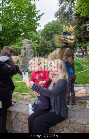 Mutter hält ihren Sohn in den Armen und nimmt ein Selfie mit ihrem Handy beim Dinosaurierfest. Stockfoto