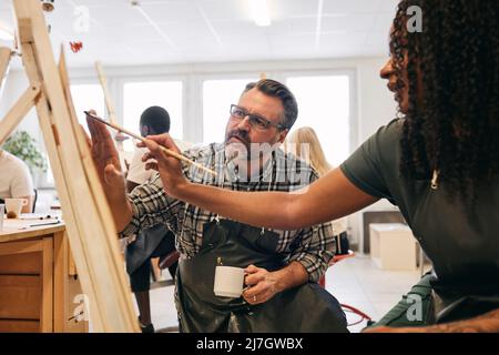 Männlicher Tutor, der weiblichen Studenten bei der Malerei auf der Leinwand des Künstlers in der Kunstklasse assistiert Stockfoto