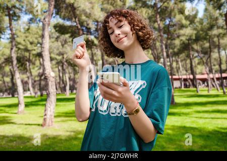 Glückliche junge Rothaarige Frau im grünen T-Shirt zeigt Plastikkreditkarte während der Verwendung von Handy. Outdoor-Online- oder Mobile-Shopping-Konzepte. Stockfoto