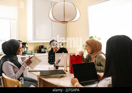 Fröhliche junge Frauen in Hijabs, die in der Küche am Tisch sitzen Stockfoto