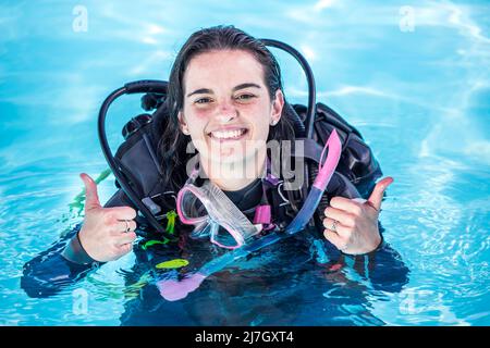 Junge Frau mit Tauchausrüstung in einem Pool lächelt an der Kamera und zeigt Daumen nach oben Stockfoto