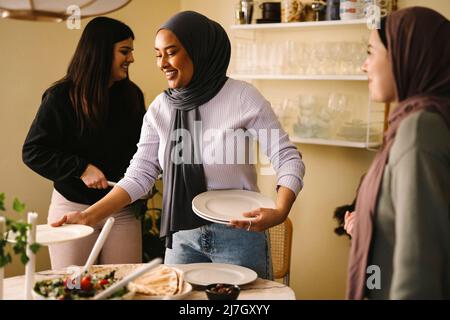 Lächelnde junge Frau, die Teller am Tisch anordnete, während sie neben Freunden in der Küche stand Stockfoto