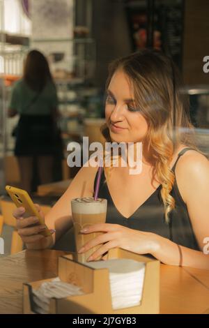 Geschäftsfrau, die in einem Café mit Kaffee sitzt, auf einem Smartphone schreibt und durch das Fenster hindurchblicken kann Stockfoto