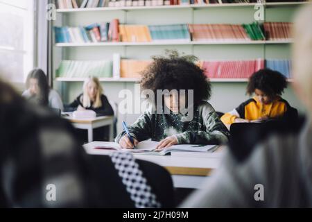 Junge mit Afro-Frisur schreibt im Buch, während sie im Klassenzimmer am Schreibtisch sitzt Stockfoto