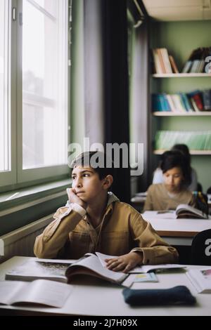 Nachdenklicher Junge mit der Hand auf dem Kinn, der im Klassenzimmer vor dem Fenster schaute Stockfoto