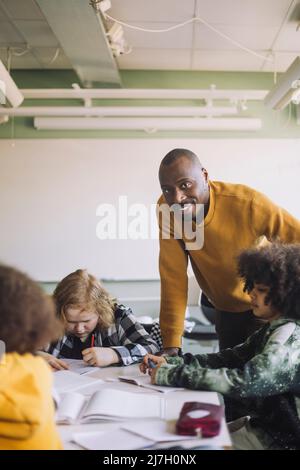 Porträt eines lächelnden männlichen Lehrers, der sich am Schreibtisch lehnt, während die Schüler im Klassenzimmer studieren Stockfoto