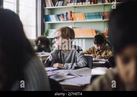 Schüler mit der Hand am Kinn, der im Klassenzimmer am Schreibtisch sitzt Stockfoto