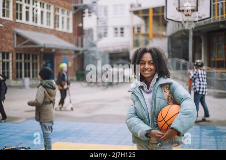 Porträt eines lächelnden Mädchens mit Basketball, das auf dem Sportplatz auf dem Schulgelände steht Stockfoto