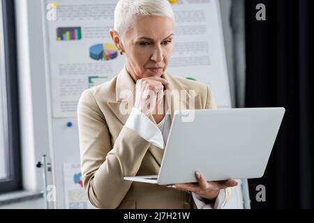 Umsichtiger Bankier, der mit einem Laptop in der Nähe des Flipchart mit Infografiken auf unscharfem Hintergrund steht Stockfoto