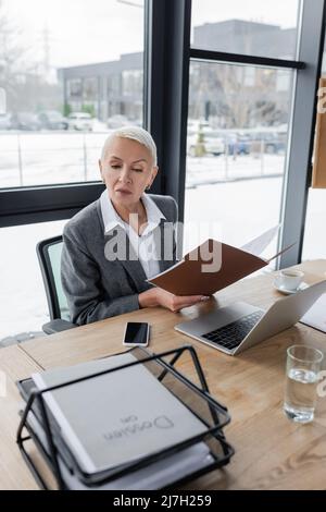 Senior Economist hält Ordner mit Dokumenten in der Nähe von Laptop und Smartphone mit leerem Bildschirm Stockfoto