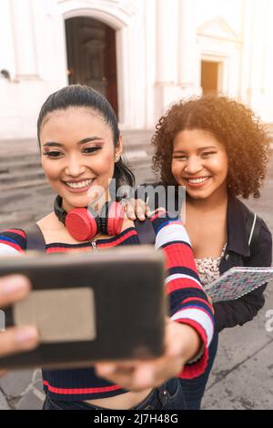 Vertikales Foto von zwei multirassischen Latina-Studenten, die auf einem öffentlichen Platz in Leon, Nicaragua, ein Selbstporträt mit einem lächelnden Handy im Freien machen. Stockfoto
