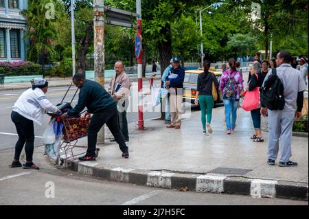 Der kubanische Mann hilft einer kubanischen Frau, ihren Einkaufswagen voller Äpfel, die sie verkauft, an die Straßenecke im Abschnitt Vedado in Havanna, Kuba, zu heben. Stockfoto