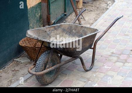 Auf dem Bürgersteig steht eine leere Schubkarre für den Garten mit einem Rad. Eine alte graue Schubkarre auf dem Gehweg auf einer Baustelle Stockfoto