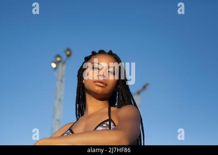 Niedriger Winkel der jungen afroamerikanischen Frau mit lockigen Haaren, die gegen den wolkenlosen blauen Himmel stehen und die Kamera betrachten Stockfoto