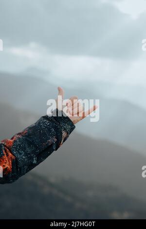Nicht erkennbare männliche Hand trägt einen Mantel, der das hängelose Zeichen auf einem Berg mit Gipfeln im Hintergrund macht Stockfoto