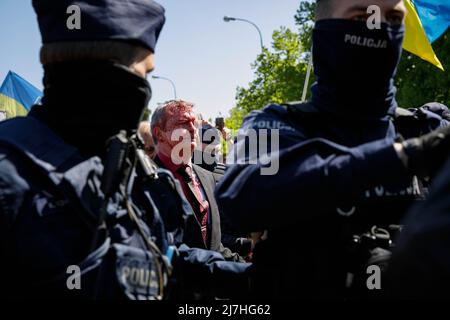 Warschau, Polen. 09.. Mai 2022. Der Botschafter der Russischen Föderation Sergej Andreew, umgeben von einem polnischen Polizeibeamten, bewegt sich in Richtung seines Autos, das mit gefälschtem Blut bedeckt ist. Am 77.. Jahrestag des Sieges der Roten Armee über Nazi-Deutschland traten ukrainische Aktivisten auf dem sowjetischen Soldatenfriedhof in Warschau auf, als sie gegen die russische Invasion in der Ukraine protestierten. Aktivisten verboten der russischen Delegation unter der Leitung von Sergej Andreev, dem Botschafter der Russischen Föderation in Polen, den Eintritt in den Friedhof. Während dieses Versuchs wurden Diplomaten von ukrainischen Aktivisten mit gefälschtem Blut übergossen. Stockfoto