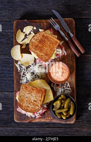 reuben Sandwich mit Roggenbrot dünn geschnittenes Corned Beef, Sauerkraut, russisches Dressing und geschmolzener Käse an Bord mit Gurken und Kartoffelchips, auf d Stockfoto