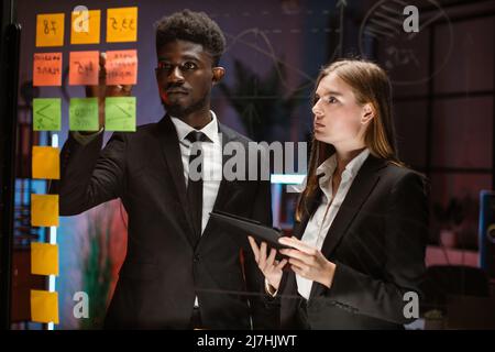 Brainstorming-Konzept. Geschäftsleute, eine hübsche kaukasische Dame und ein afroamerikanischer Mann treffen sich abends im modernen Büro und nutzen Post-IT-Notizen, um Ideen auszutauschen. Haftnotiz auf Glaswandtafel. Stockfoto