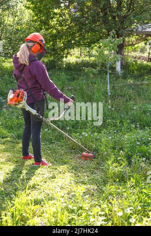 Die Frau mäht das Gras im Garten mit einem Benzintrimmer. Sie trägt eine Brille und einen Helm mit Kopfhörern. Arbeiten Sie im Frühling im Garten. Stockfoto