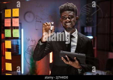 Vorderansicht des jungen afroamerikanischen Geschäftsmannes, der im Büro auf einer Glasscheibe sprach und schrieb. Blick durch die Glastafel mit bunten Haftnotizen für die Planung oder die Projektphasen Stockfoto