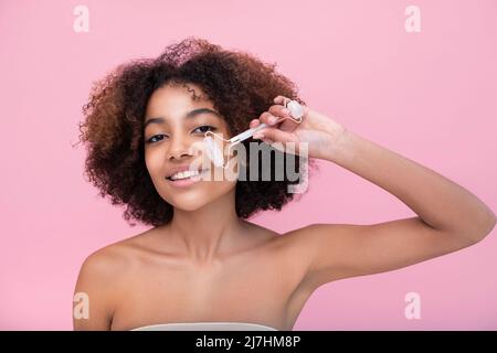 Porträt einer jungen, gepflegten dunkelhäutigen Frau mit üppigen Haaren, die mit Hilfe einer Walze mit geschlossenen Augen auf einem rosa Hintergrund Nahaufnahme, Pflege und Hautpflege eine Gesichtsmassage macht Stockfoto